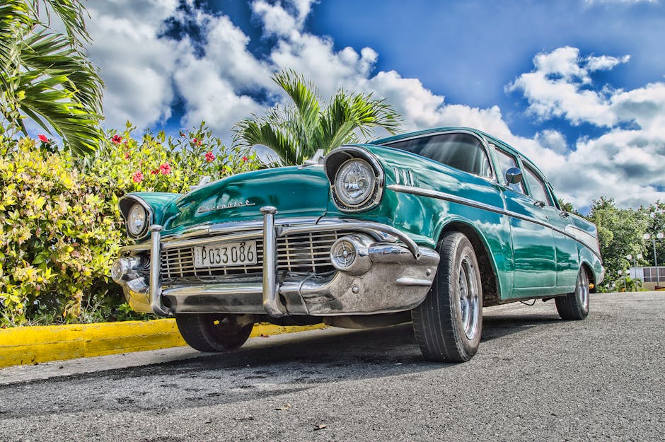 A beautifully restored classic car on a sunny road in Havana, Cuba, with lush foliage and a vibrant sky.